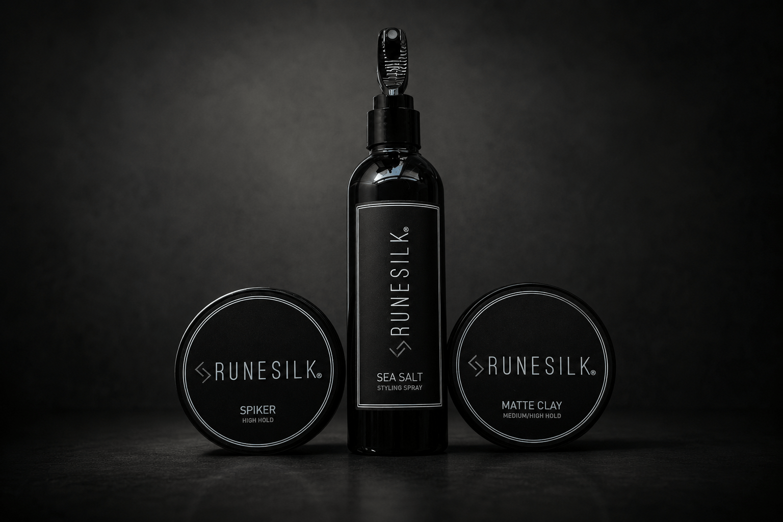Three black cosmetic containers from the brand RUNESILK are arranged on a dark surface against a black background. The center is a tall bottle labeled SEA SALT, flanked by two round jars labeled SPYDER and MATTE CLAY.