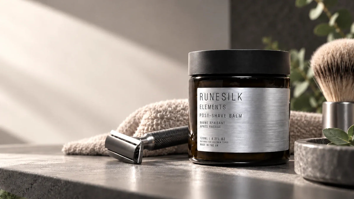 A jar of Runesilk Elements Post-Shave Balm stands on a stone surface with a safety razor, towel, and shaving brush in a softly lit bathroom-like setting.